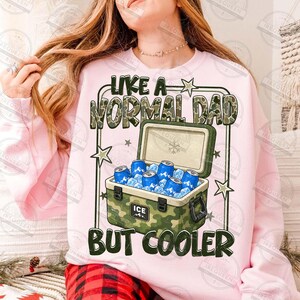 May include: A light pink sweatshirt with the text "LIKE A NORMAL DAD BUT COOLER" and a graphic of a camouflage cooler filled with ice and blue cans. The cooler has the word "ICE" on it.