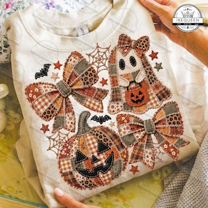 May include: A cream-colored sweatshirt featuring a patchwork-style graphic print of a jack-o'-lantern, a ghost holding a pumpkin bucket, and two large bows. The design includes orange, brown, and black patterns, spiderwebs, bats, and stars.