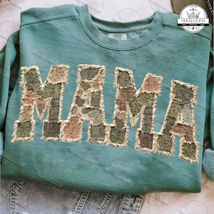 May include: A teal sweatshirt featuring the word "MAMA" in a patchwork design. The letters are crafted from various camouflage fabrics in green, brown, and tan hues, with a distressed, frayed edge.