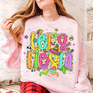May include: A pale pink sweatshirt featuring the phrase "Let's Fiesta" in vibrant, decorative lettering. The design incorporates a sombrero, cactus, taco, and other celebratory elements, ideal for a party.