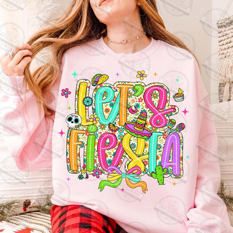 May include: A pale pink sweatshirt featuring the phrase "Let's Fiesta" in vibrant, decorative lettering. The design incorporates a sombrero, cactus, taco, and other celebratory elements, ideal for a party.