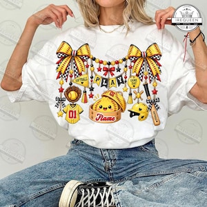 Może przedstawiać: Biały t-shirt z kolorową grafiką o tematyce softballowej. Wzór zawiera żółte kokardy, rękawicę do softballu, koszulkę z numerem 01, kij, kask oraz postać pierożka w koszyku do gotowania na parze z napisem Name.