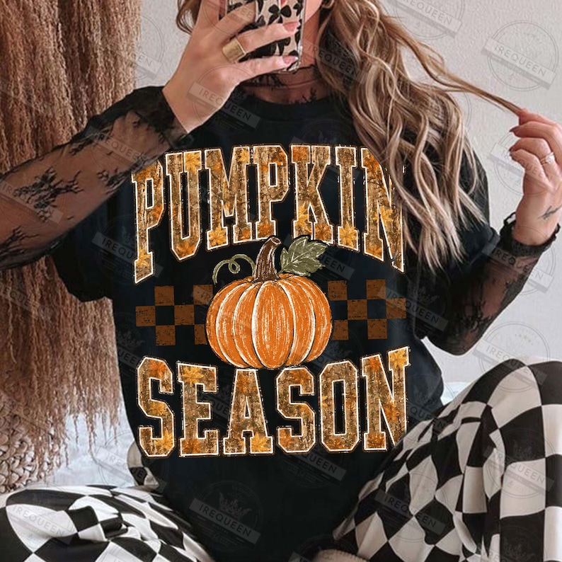 May include: A black short-sleeved t-shirt featuring the text PUMPKIN SEASON in a block font with a fall leaf pattern. A large orange pumpkin graphic is centered between the words. A brown and black checkered pattern sits behind the pumpkin.