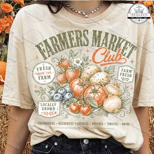 May include: A cream-colored t-shirt featuring a graphic print with the text Farmers Market Club. The design includes illustrations of strawberries, blueberries, cherries, potatoes, tomatoes, and onions, along with the phrases Fresh From The Farm and Locally Grown.