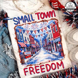 Könnte beinhalten: Weißes T-Shirt mit einem patriotischen Design, das eine Kleinstadt-Straßenszene in Rot, Weiß und Blau zeigt. Die Worte "SMALL TOWN FREEDOM" sind deutlich zu sehen. Das Shirt ist mit einer silbernen Kette verziert.