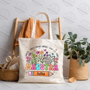 May include: A white canvas tote bag with a colorful floral design and the word "MAESTRA" in large, colorful letters. The name "Jullia" is written below the word "MAESTRA" in a stylized font. A pencil with a pink eraser is drawn next to the name. The bag has a circle of words around the floral design, including "compasivo", "cari&ntilde;osa", "dedicada", "confiable", "c&aacute;lida", "leal", and "amable".