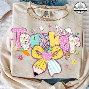 May include: A beige sweatshirt with the word "Teacher" in colorful, patterned letters. The design includes a carrot, an apple, a pencil, and bunny ears, with a gold chain necklace. The sweatshirt is a Comfort Colors brand.