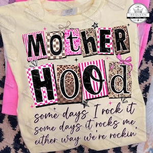 Può includere: Una felpa giallo chiaro con la scritta "MOTHERHOOD" in un design a blocchi con motivi rosa, marroni e neri. Il testo "some days I rock it, some days it rocks me, either way we're rockin'" è stampato sotto.