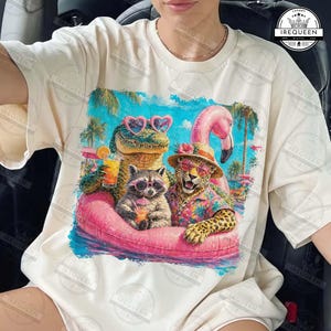 May include: Cream-colored t-shirt featuring a vibrant graphic of a crocodile, raccoon, and leopard in sunglasses, lounging on a pink inflatable ring. The design includes a flamingo, tropical drinks, and palm trees, creating a fun, vacation-themed look.