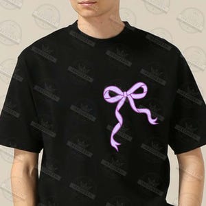 May include: Black t-shirt featuring a light purple bow design on the front. The bow has flowing ribbons and is centered on the chest. The shirt is short-sleeved and made of a soft material.