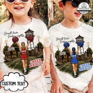 Op de afbeelding: Twee off-white t-shirts met een afbeelding van een cheerleader op een voetbalveld met een watertoren, de tekst Small town Friday nights en Cheer Mascot. Het linker t-shirt heeft een rood uniform, het rechter een blauw uniform.