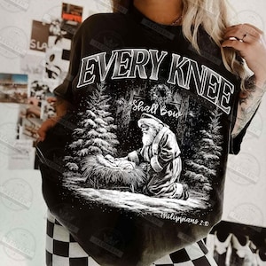 May include: Black t-shirt with a graphic featuring Santa kneeling before baby Jesus in a snowy forest scene. The text "EVERY KNEE Shall Bow" is arched above the image, with "Philippians 2:10" below.