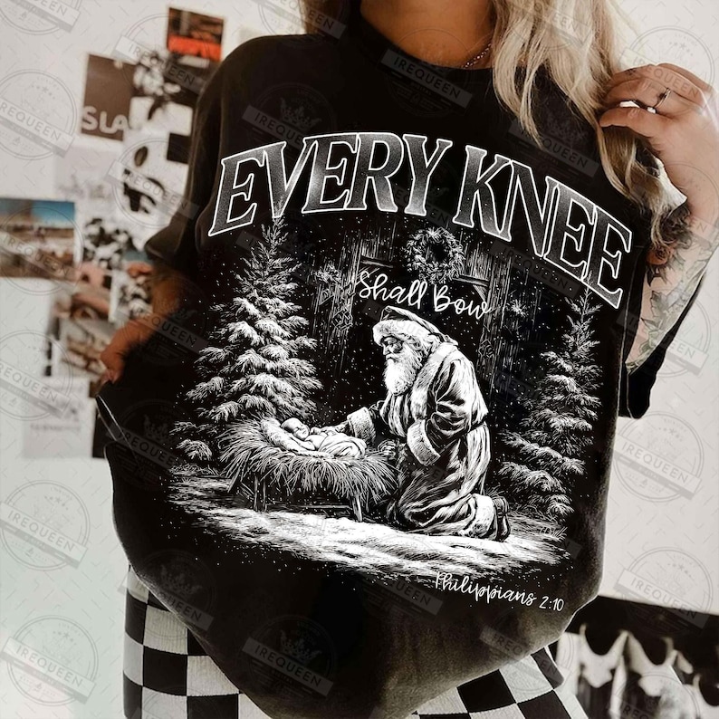 May include: Black t-shirt with a graphic featuring Santa kneeling before baby Jesus in a snowy forest scene. The text "EVERY KNEE Shall Bow" is arched above the image, with "Philippians 2:10" below.