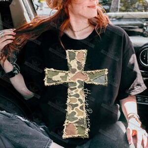 May include: Black t-shirt featuring a camouflage cross design with frayed edges. The cross is made of various shades of green, brown, and tan. The words "He is Risen" are written vertically on the right side of the cross in a script font. The shirt is a casual style.
