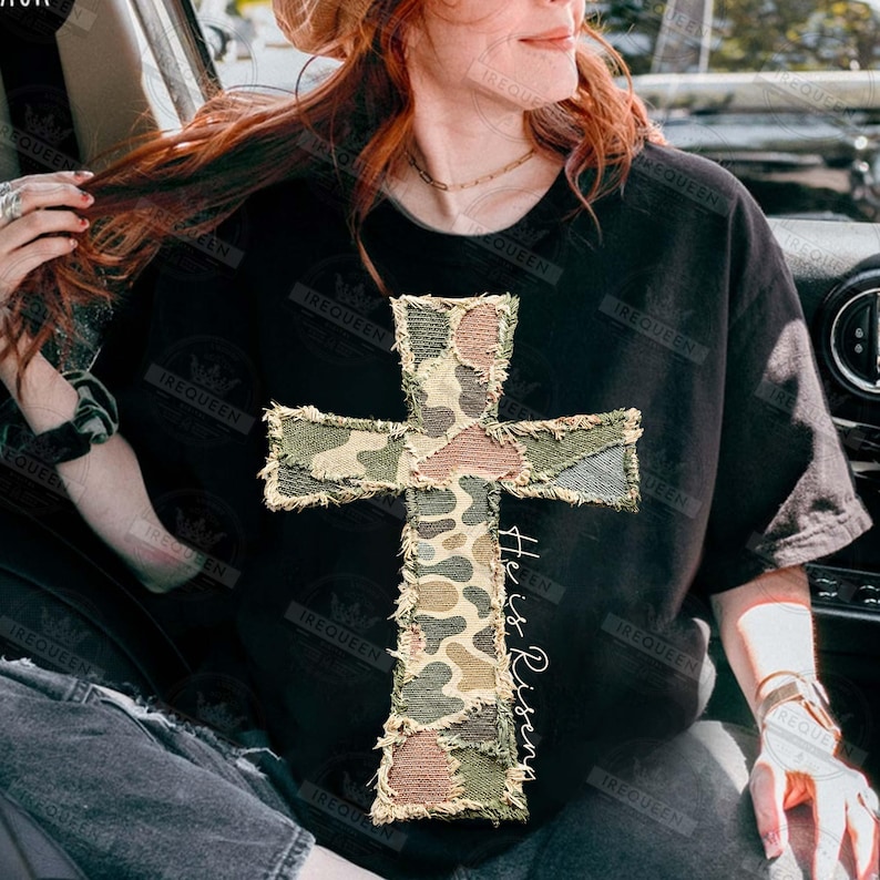 May include: Black t-shirt featuring a camouflage cross design with frayed edges. The cross is made of various shades of green, brown, and tan. The words "He is Risen" are written vertically on the right side of the cross in a script font. The shirt is a casual style.