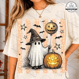 May include: A cream-colored t-shirt featuring a graphic of a ghost wearing a witch hat, holding a pumpkin balloon and standing next to a jack-o'-lantern. The design includes orange vertical stripes, stars, and bats on a light background.