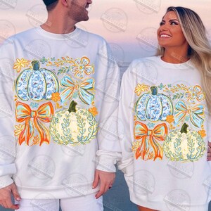 May include: White sweatshirts with fall-themed designs. Each sweatshirt features a pumpkin with a blue floral pattern, a bow, and a pumpkin with a leafy design. The designs also include orange and yellow floral accents.