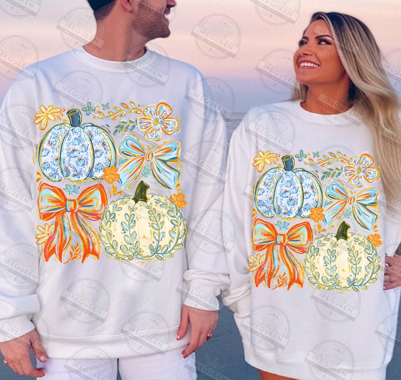 May include: White sweatshirts with fall-themed designs. Each sweatshirt features a pumpkin with a blue floral pattern, a bow, and a pumpkin with a leafy design. The designs also include orange and yellow floral accents.