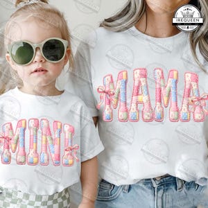 May include: Two white t-shirts, one with "MAMA" and the other with "MINI" in a patchwork design. The letters are in pastel colours with pink bows. The child wearing the "MINI" shirt is wearing sunglasses.