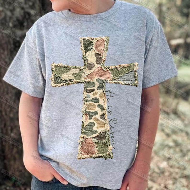 May include: A light gray t-shirt featuring a camouflage cross design with frayed edges. The cross is made of various shades of green, brown, and tan. The words "He is Risen" are written vertically on the right side of the cross.