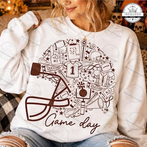 May include: A white long-sleeve sweatshirt featuring a large brown graphic print of a football helmet filled with various American football icons, including a jersey with the number 1, a football, a megaphone, and the text Game day.
