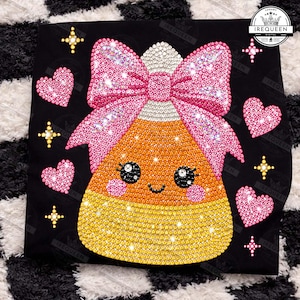 May include: A black t-shirt featuring a rhinestone-embellished candy corn character with a large pink bow, pink hearts, and yellow starbursts. The candy corn has a smiling face with black eyes and pink cheeks. The IREQUEEN logo is in the corner.