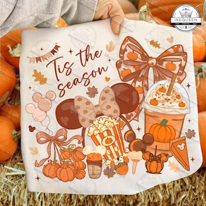 以下が含まれることがあります： 「Tis the season」という文字が書かれた、茶色とオレンジ色のグラフィックがプリントされた白いTシャツ。カボチャ、大きなリボン、ポップコーンバケツ、ドリンクカップ、ネズミの耳のシルエットが秋をテーマに描かれています。