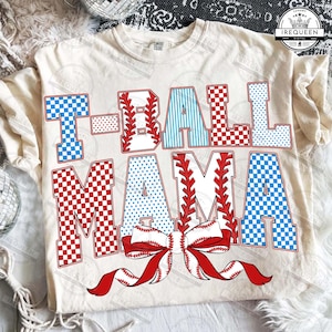 Puede incluir: Camiseta color crema con el texto "T-BALL MAMA" en letras de bloque rojas, blancas y azules. Las letras tienen patrones temáticos de béisbol y un lazo de béisbol en la parte inferior. La camiseta es de ajuste cómodo.