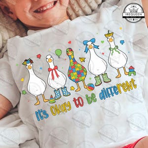 May include: White t-shirt featuring five cartoon geese with various accessories and patterns. The geese are wearing boots, bows, and flower crowns. The text "It's Okay to be different" is written in colorful letters below the geese.
