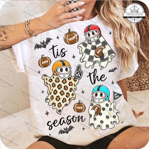 May include: A white t-shirt featuring a graphic print of three ghosts wearing football helmets, surrounded by pumpkins, bats, and stars. The ghosts have patterns including black and white checks, leopard print, and black polka dots. Text reads: tis the season.