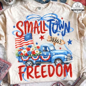 Könnte beinhalten: Cremefarbenes T-Shirt mit patriotischem Design. Es zeigt einen blauen Pickup-Truck voller Blumen, eine amerikanische Flagge und die Worte "Small Town Freedom" in Rot und Blau. Enthält die Nummer 38863.