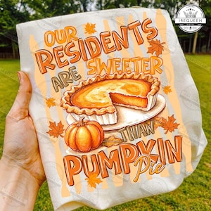 May include: A white t-shirt featuring a graphic print of a pumpkin pie and a whole pumpkin, surrounded by orange maple leaves. The text reads, Our residents are sweeter than pumpkin pie, in orange and brown lettering with a crown logo.