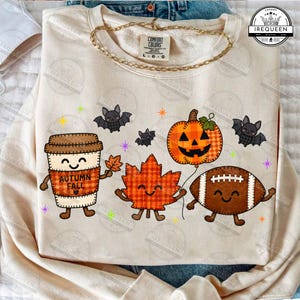 May include: A beige crewneck sweatshirt featuring a graphic print of a smiling coffee cup with the text AUTUMN FALL, a plaid pumpkin, a plaid maple leaf, and a brown football, all with arms and legs, surrounded by small bats and stars.
