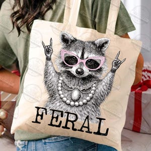 Puede incluir: Bolso tote beige con un mapache con gafas de sol rosas y un collar de perlas, con la palabra "FERAL" en letras negras en negrita. El mapache hace un gesto de rock and roll con la mano. La bolsa es de tela clara.