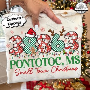 Może przedstawiać: Kremowa bluza z numerami "3863" w świątecznym wzorze. Numery są ozdobione czapkami Świętego Mikołaja, rogami reniferów i bałwanem. Tekst "PONTOTOC, MS Small Town Christmas" jest wydrukowany poniżej.