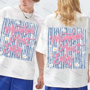May include: White t-shirts with the text "Mommin' Ain't Easy" in pink script, set against a blue striped background, adorned with daisy accents. The shirts are short-sleeved and have a casual fit.