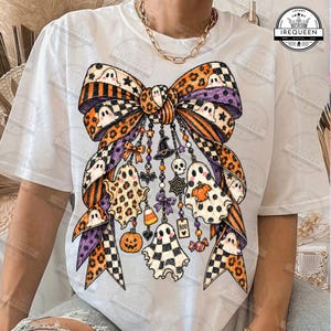 May include: A white t-shirt featuring a large graphic of a decorative bow with a Halloween theme. The bow pattern includes orange and black stripes, leopard print, and checkered squares. Hanging from the bow are charms of ghosts, pumpkins, a witch hat, a skull, a spider web, candy corn, and a tag with the text BOO.