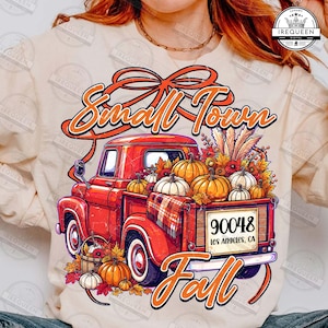May include: A cream-colored long-sleeve shirt featuring a graphic of a red vintage pickup truck filled with pumpkins and autumn foliage. The text reads Small Town Fall and 90048 Los Angeles, CA. The design includes a plaid blanket and a basket.
