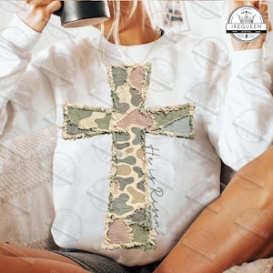 May include: White sweatshirt featuring a large, textured cross applique with a camouflage pattern in shades of green, brown, and pink. The words "He is Risen" are written vertically on the right side of the cross.