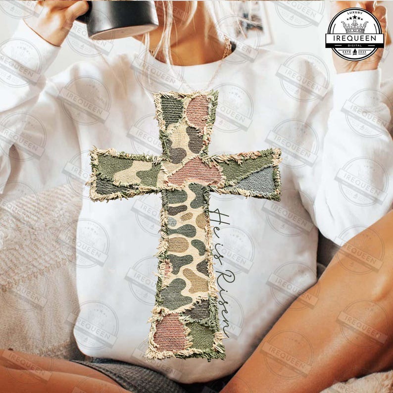 May include: White sweatshirt featuring a large, textured cross applique with a camouflage pattern in shades of green, brown, and pink. The words "He is Risen" are written vertically on the right side of the cross.