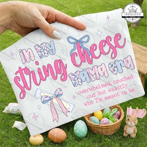 Könnte beinhalten: Eine weiße Decke mit dem Text "In My String Cheese Mama Era" in rosa und blauen Buchstaben. Die Decke hat ein florales Bogendesign und ist von Osterdekorationen umgeben, darunter Eier und ein Hase.