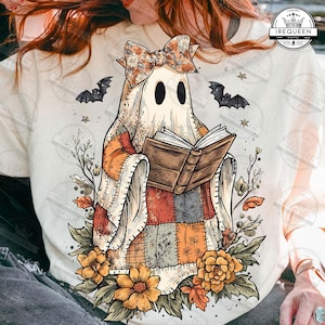 May include: A cream-colored t-shirt featuring a whimsical ghost reading a book. The ghost is adorned with a floral bow and patchwork quilt-like design. Bats and stars surround the ghost, with fall foliage and flowers at the base.