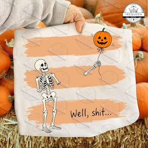 May include: A white t-shirt featuring a graphic of a skeleton holding a string attached to a jack-o'-lantern pumpkin balloon. The design includes three horizontal orange brushstroke stripes and the black text Well, shit... printed below.