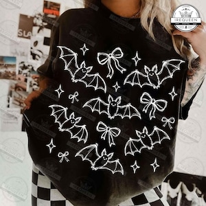 May include: A black t-shirt featuring a white graphic print of five bats, four bows, and several four-pointed stars scattered across the front. The design is arranged in a repeating pattern, showcasing the bats in various flying positions.