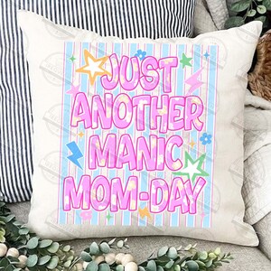 May include: A white square pillow with the text "JUST ANOTHER MANIC MOM-DAY" in pink, outlined in a lighter pink. The text is set against a light blue and white striped background, with star and lightning bolt graphics.