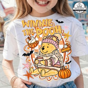 May include: A white t-shirt featuring a graphic of Winnie the Pooh wearing a pink knit hat and scarf, holding a coffee cup. The design includes orange pumpkins, a slice of pie, black bats, and pink and orange stars with the text WINNIE THE POOH.