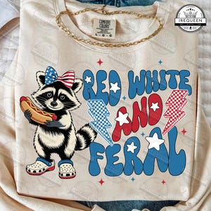 May include: Cream-colored sweatshirt with a raccoon graphic wearing a patriotic bow and holding a hot dog. The text reads "RED WHITE AND FERAL" in red and blue. Includes a gold chain necklace.