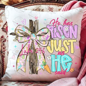 May include: White square pillow with a colorful Easter design. The design includes a wooden cross with a pink bow adorned with bunnies and flowers. Text reads "He has risen just as He said" and "Luke 24:6".