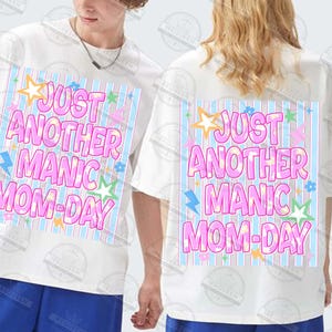 May include: White t-shirts with the text "JUST ANOTHER MANIC MOM-DAY" in pink, blue, and yellow. The design includes stars, lightning bolts, and flowers. The shirts are short-sleeved.