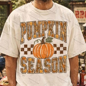 May include: A cream-colored short-sleeve t-shirt featuring a graphic print with the text PUMPKIN SEASON in a brown wood-grain pattern. Centered between the words is a large orange pumpkin with green leaves and a brown checkered border.